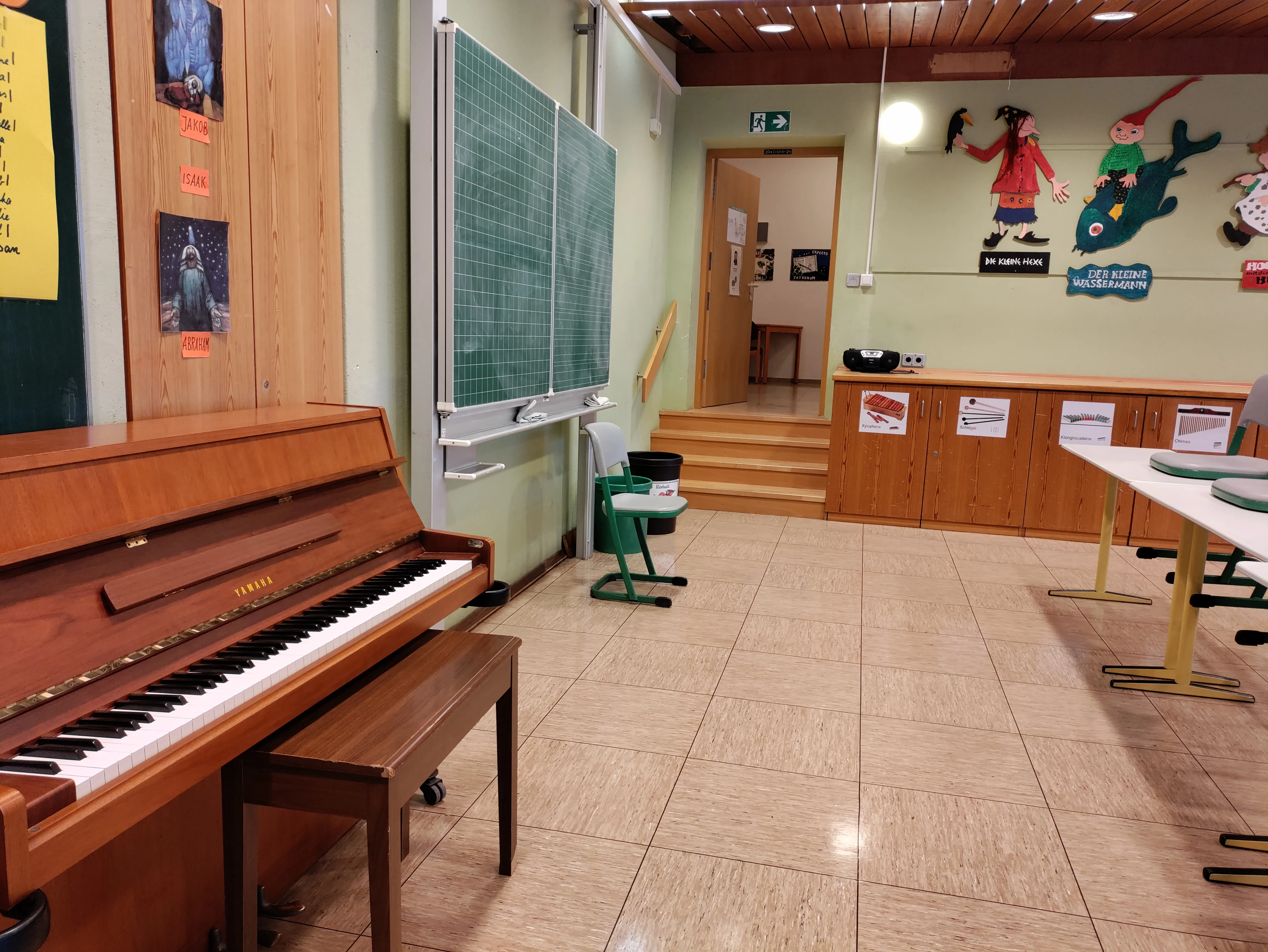 Ein Klavier aus hellem Holz in der Aula, an der Wand dahinter Bilder der Kinder.