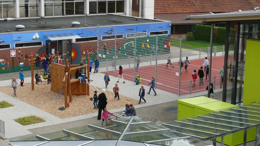 Blick von oben auf den Schulhof mit Kletterturm, Sportfeld und spielenden Kindern.